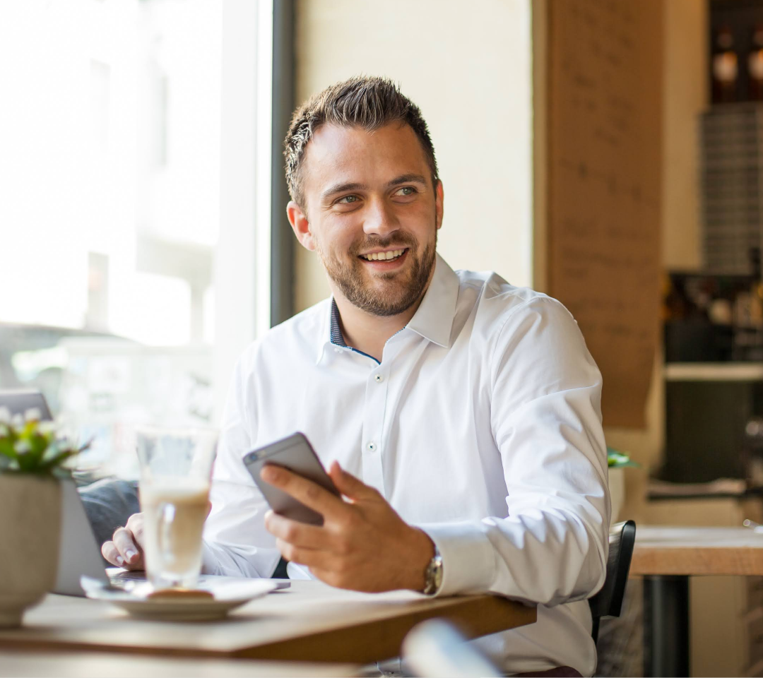 Daniel Palm lacht und sitzt an einem Tisch. Dabei hält ein Iphone in der Hand. Auf dem Tisch steht ein Kaffee und ein geöffneter Laptop. 