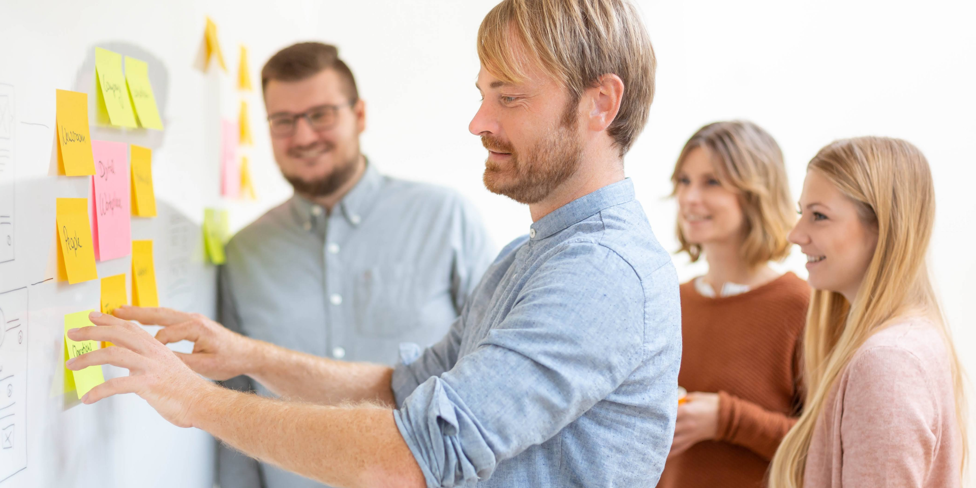 Zwei Männer und zwei Frauen stehen vor einem Whiteboard und kleben bunte Post-its auf