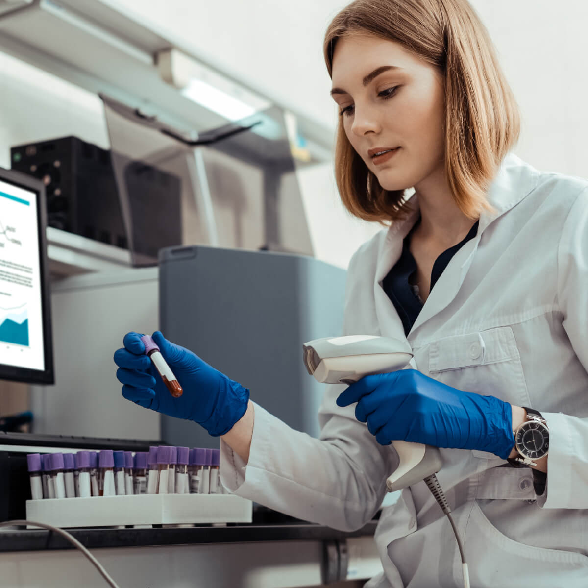 a woman in a lab coat and blue gloves, working in a laboratory setting. She is holding a blood sample tube in one hand and a barcode scanner in the other. The computer screen in the background displays a graph or data, indicating the scientific context of the work. The setting appears clean and professional, typical of a laboratory environment focused on testing or research | AI Agency SUNZINET