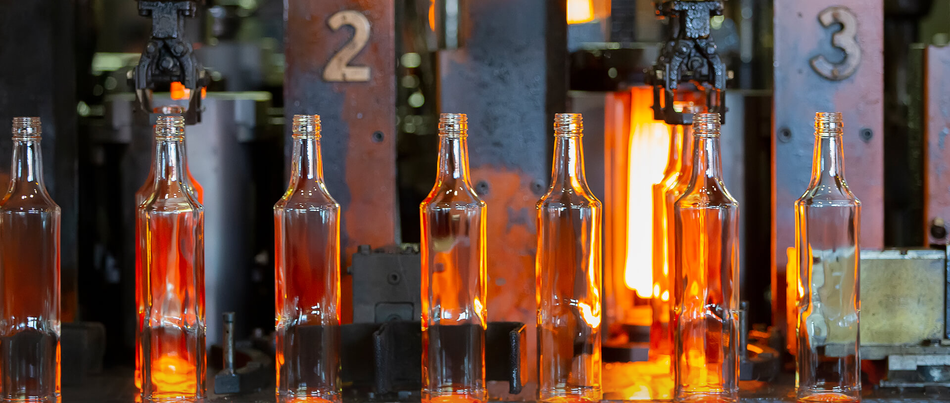 A close-up view of glass bottles being manufactured in a factory. The bottles are still glowing orange from the heat and are in the process of being molded by machines, with numbers visible on the machinery.