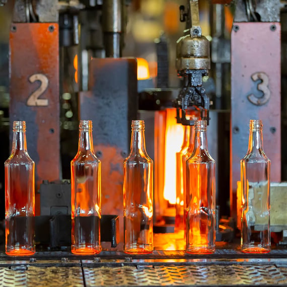 Eine Nahaufnahme von Glasflaschen, die in einer Fabrik hergestellt werden. Die Flaschen leuchten noch orange vor Hitze und werden gerade von Maschinen geformt, wobei auf den Maschinen Nummern zu sehen sind.
