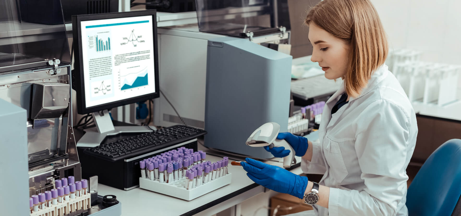 Die Person im weißen Labormantel scannt mit einem Barcode-Scanner eine Probe, die sie in der Hand hält. Vor ihr steht ein Testtubengestell mit Vials, während auf dem Computerbildschirm im Hintergrund Diagramme und Daten sichtbar sind.