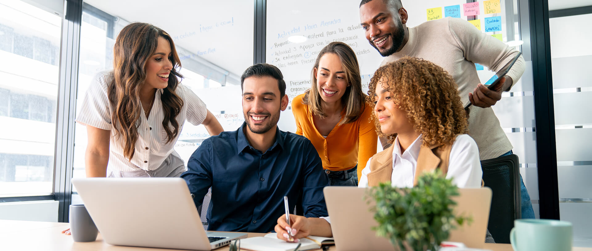The photo shows a group of five colleagues in a modern office setting, gathered around a table and collaborating on a project. They are smiling and engaging with each other, looking at a laptop screen. Two people are sitting, with one holding a pen, while three are standing behind them. There are colorful sticky notes on the glass wall in the background, and a plant is placed on the table. The atmosphere is dynamic and collaborative.
