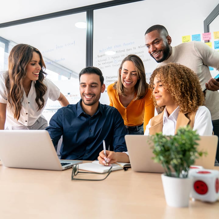 Das Foto zeigt fünf Kollegen in einem modernen Büro. Sie sitzen um einen Tisch und arbeiten gemeinsam an einem Projekt. Sie lächeln, unterhalten sich angeregt und blicken auf einen Laptop-Bildschirm. Zwei Personen sitzen, eine hält einen Stift, während drei hinter ihnen stehen. An der Glaswand im Hintergrund kleben bunte Haftnotizen, und auf dem Tisch steht eine Pflanze. Die Atmosphäre ist dynamisch und kooperativ.