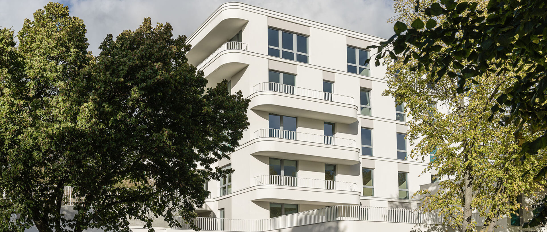 Modern white apartment building with rounded balconies and large windows, partially framed and obscured by leafy trees in daylight.