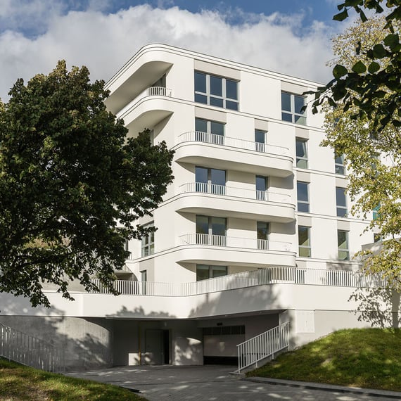 Modern white apartment building with rounded balconies and large windows, partially framed and obscured by leafy trees in daylight.