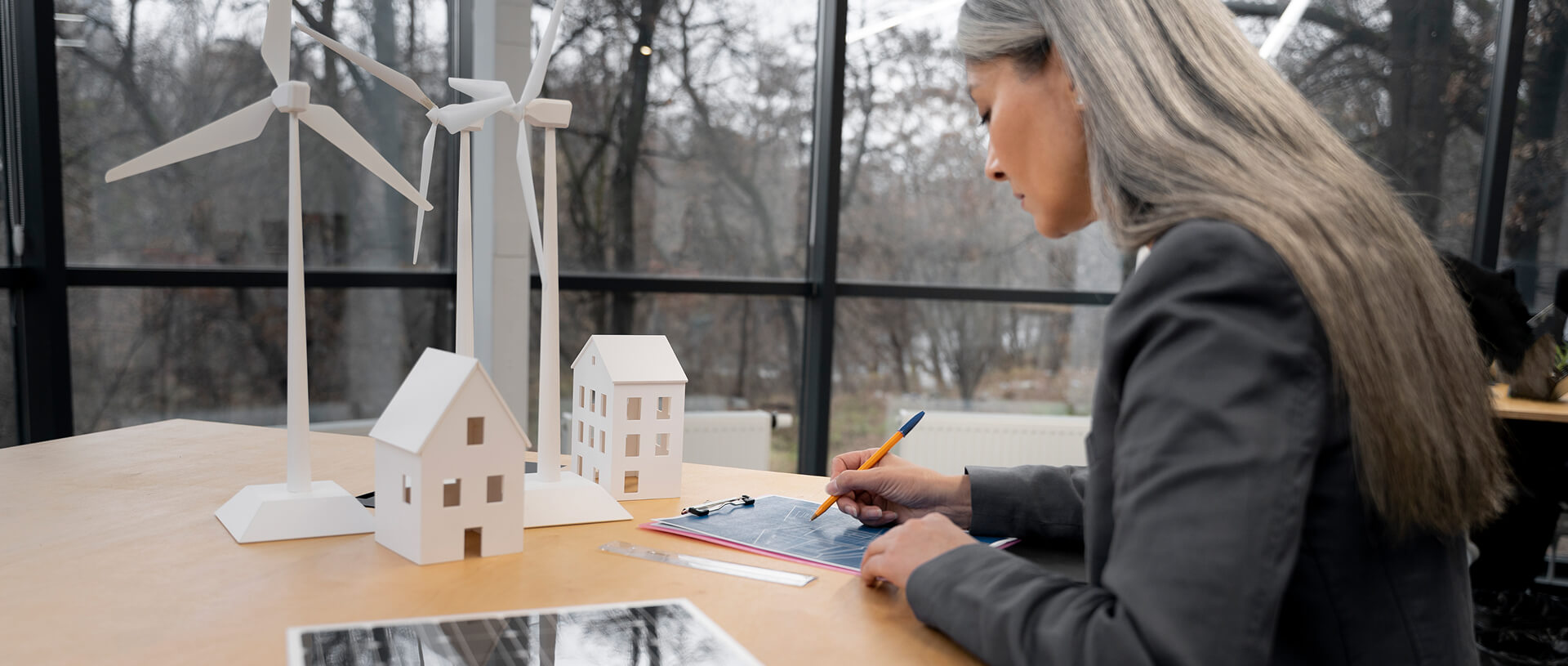 A woman with long gray hair is sitting at a desk, working on a blueprint with a pencil. The desk features architectural models of houses, a solar panel, and wind turbines, with large windows showing a natural landscape in the background. | legacy system modernization SUNZINET