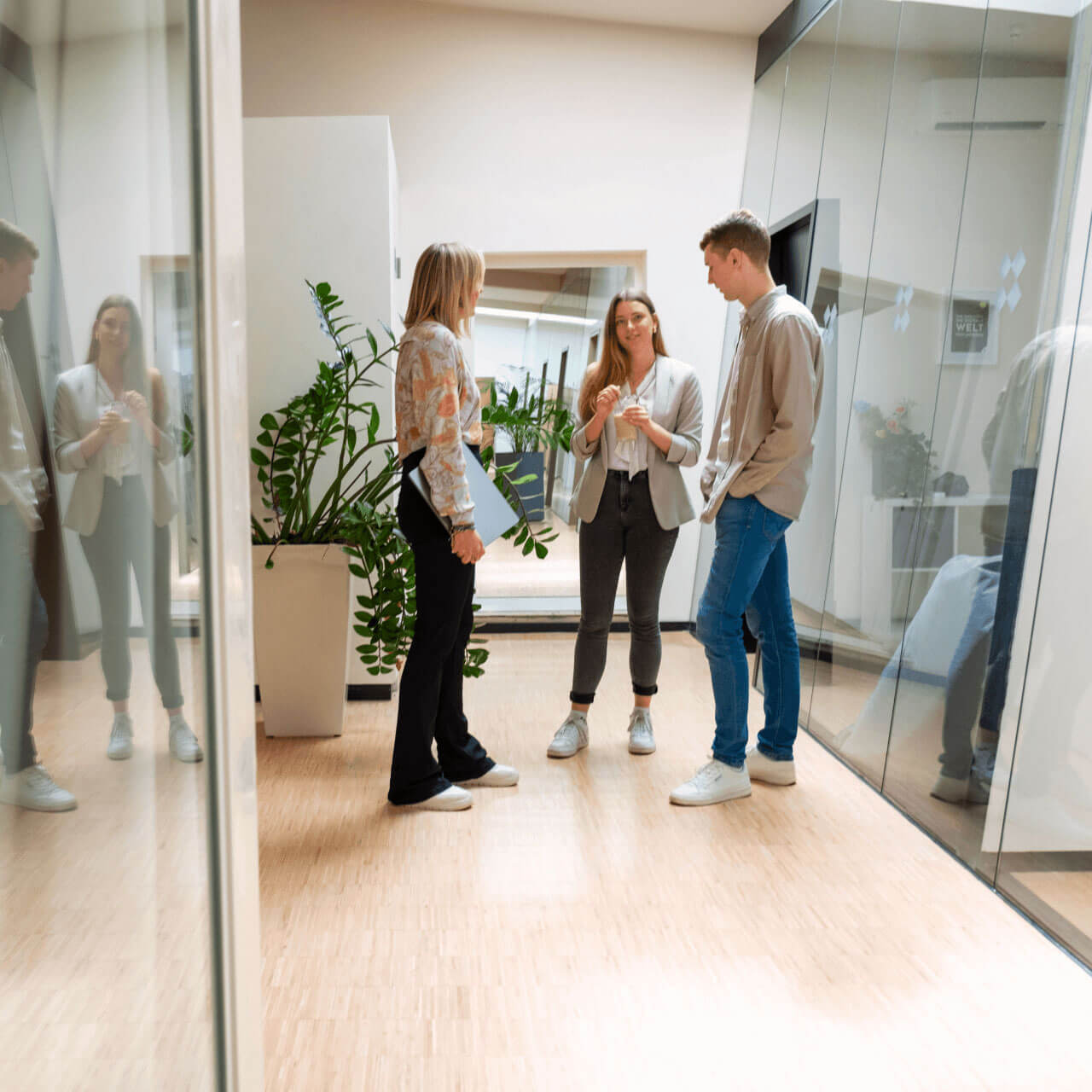 Drei junge Menschen unterhalten sich ungezwungen in einer modernen Büroumgebung. Zwei von ihnen, eine Frau und ein Mann, stehen in der Nähe einer Glaswand, umgeben von großen Topfpflanzen. Die Frau hält einen Laptop in der Hand, der Mann ist leger gekleidet und trägt ein beiges Hemd und Blue Jeans. Die Frau in der Mitte hat langes Haar und trägt eine helle Bluse und eine schwarze Hose.