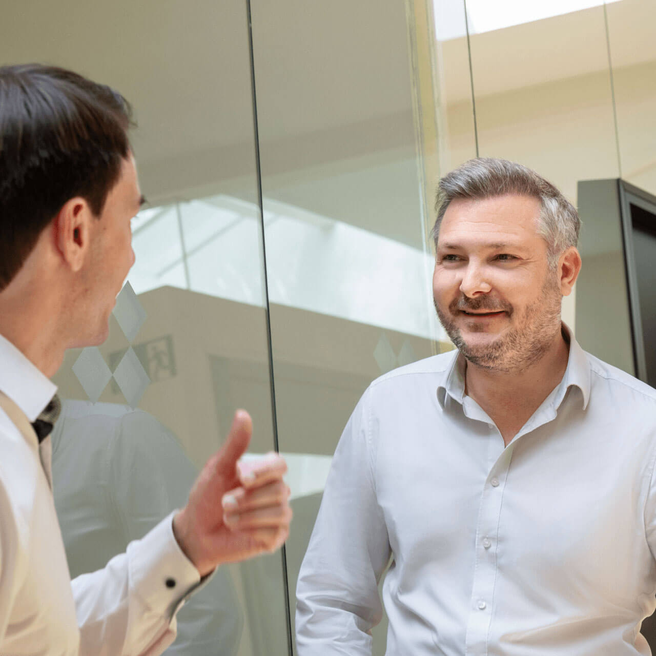 Zwei Männer in einer professionellen Umgebung unterhalten sich. Der Mann links trägt ein weißes Hemd und gestikuliert mit der Hand, wobei er den Daumen nach oben streckt. Der andere Mann rechts lächelt und trägt ein hellgraues Hemd. Im Hintergrund ist eine Glastrennwand zu sehen, die eine moderne Büroatmosphäre schafft.