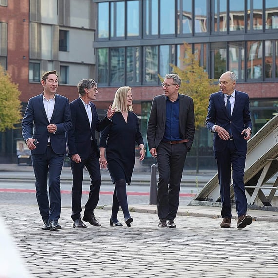 Eine Gruppe von fünf Anwälten in Anzügen geht einen gepflasterten Weg entlang. Hinter ihnen erhebt sich ein modernes Bürogebäude. Sie sind alle in ein Gespräch vertieft.