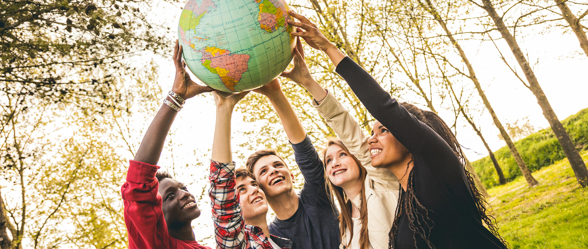 eine Gruppe von fünf verschiedenen jungen Menschen, die im Freien stehen und gemeinsam eine Weltkugel halten. Sie alle lächeln und heben die Weltkugel mit beiden Händen über ihre Köpfe. Im Hintergrund sind Bäume mit grünen Blättern zu sehen, und die Gesamtatmosphäre wirkt positiv und geeint. | content optimierung SUNZINET