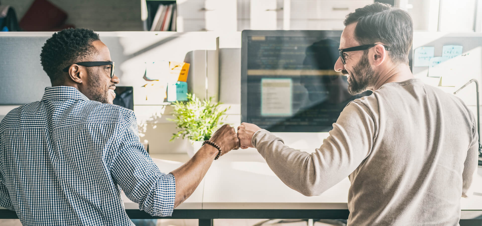 Zwei Männer mit Brille sitzen nebeneinander an Schreibtischen in einem hellen, modernen Büro. Sie lächeln und geben sich einen Fist Bump – ein Moment der Freude oder des Teamgeists.