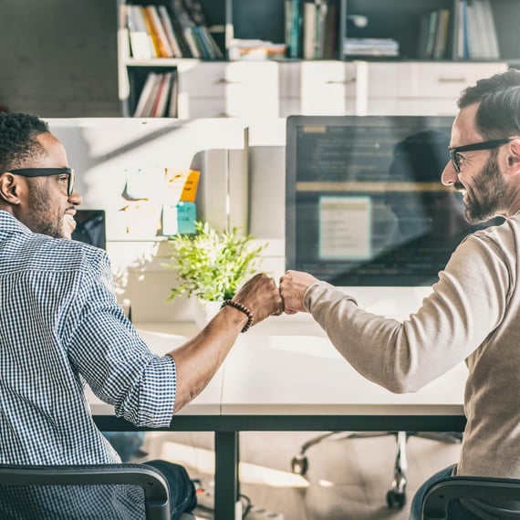 Zwei Männer mit Brille sitzen nebeneinander an Schreibtischen in einem hellen, modernen Büro. Sie lächeln und geben sich einen Fist Bump – ein Moment der Freude oder des Teamgeists.