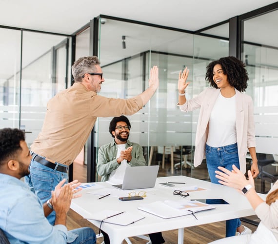 eine Gruppe von Kollegen in einer modernen Büroumgebung, die einen Erfolg feiern. Ein Mann gibt einer Frau ein High-Five, während zwei andere im Hintergrund klatschen und lächeln. | innovationskultur SUNZINET
