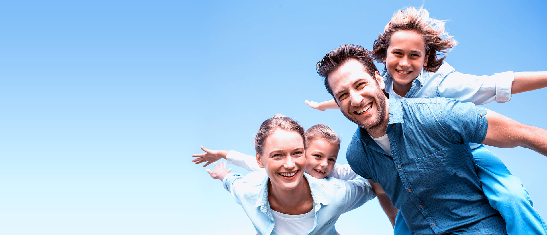 Lächelnde vierköpfige Familie draußen unter klarem blauem Himmel, zwei Erwachsene tragen ihre Kinder auf dem Rücken, während alle spielerisch die Arme ausstrecken, als würden sie fliegen.