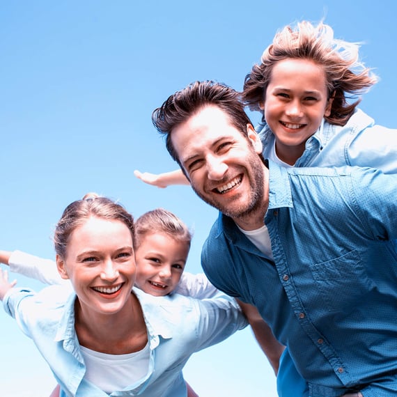 Lächelnde vierköpfige Familie draußen unter klarem blauem Himmel, zwei Erwachsene tragen ihre Kinder auf dem Rücken, während alle spielerisch die Arme ausstrecken, als würden sie fliegen.