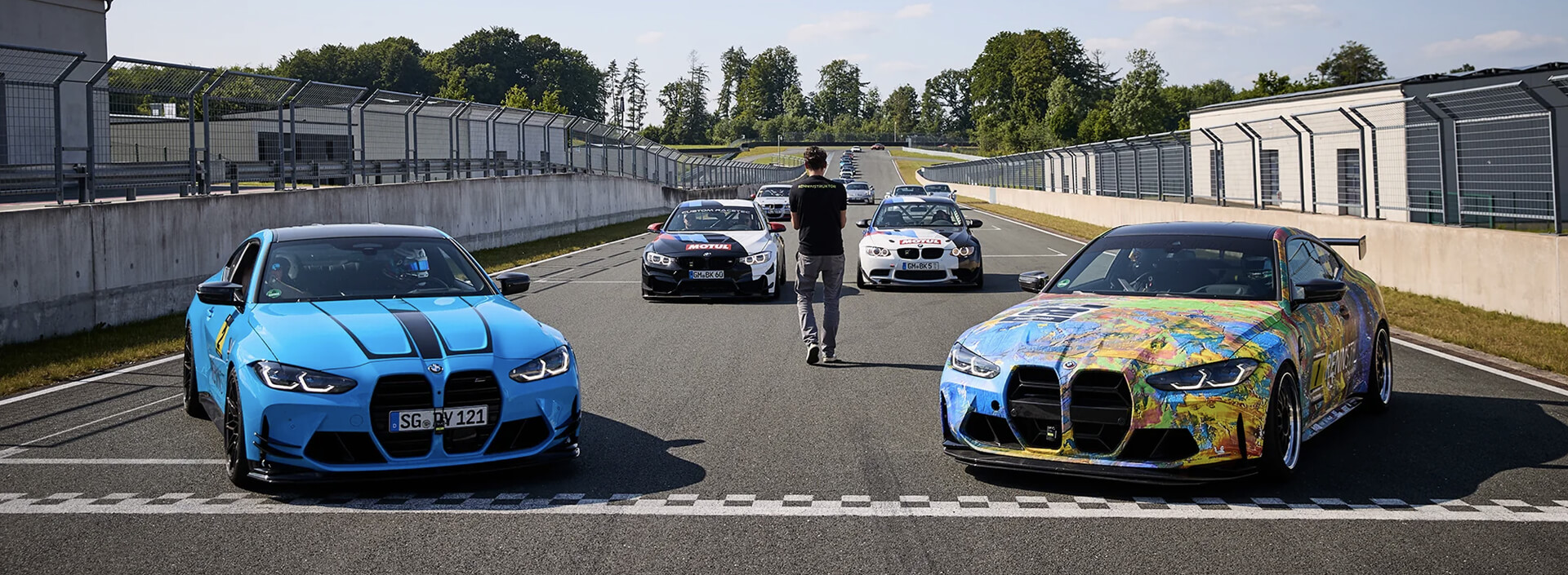 Several BMW sports cars lined up on a racetrack starting grid, with two prominently in the foreground (one blue, one with a multicolored paint design), additional cars and a person standing in the center lane behind them, pit buildings and fencing on both sides under clear daylight. | Digital Agency SUNZINET