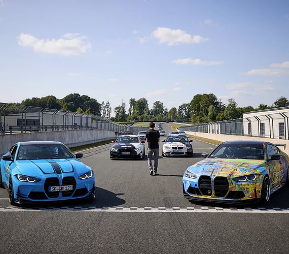 Mehrere BMW-Sportwagen stehen an der Startlinie einer Rennstrecke aufgereiht, wobei zwei davon deutlich im Vordergrund zu sehen sind (einer blau, einer mit mehrfarbiger Lackierung); weitere Fahrzeuge und eine Person stehen hinter ihnen auf der mittleren Fahrspur, an beiden Seiten sind Boxengebäude und Zäune zu sehen – alles bei strahlendem Tageslicht. | Digitalagentur SUNZINET