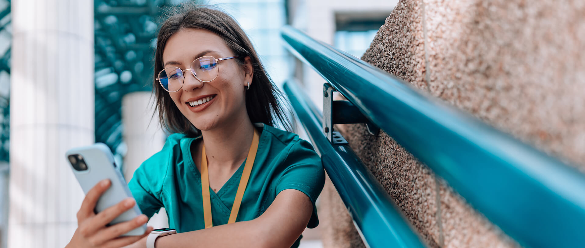 Photo showcasing a young nurse sitting outside the health clinic on the stairs. She is wearing glasses and holding a smartphone. She is wearing a green uniform.