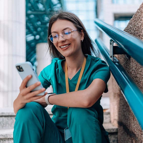 Foto, das eine junge Krankenschwester zeigt, die vor der Gesundheitsklinik auf der Treppe sitzt. Sie trägt eine Brille und hält ein Smartphone in der Hand. Sie trägt eine grüne Uniform.