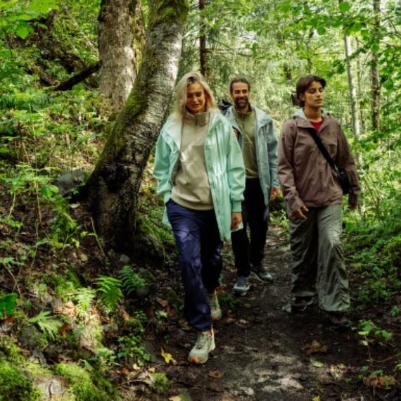 A group of three adults in going down a forest path. Two young women are in the frontand a man is following them. They are wearing light and colorful outdoor clothes.