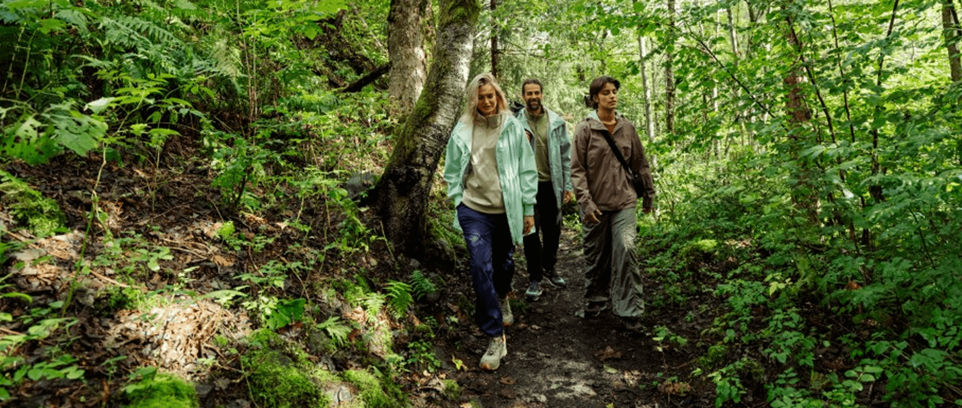 A group of three adults in going down a forest path. Two young women are in the frontand a man is following them. They are wearing light and colorful outdoor clothes.