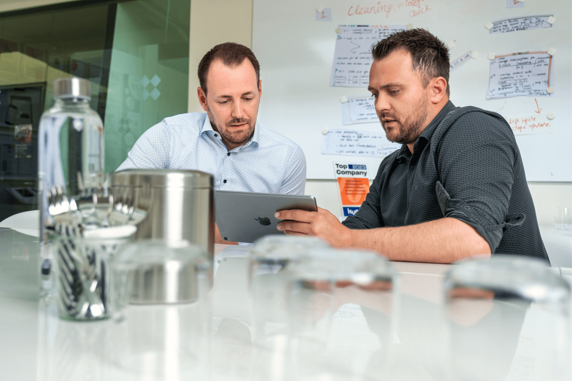 Zwei Kollegen in einem Besprechungsraum, die Inhalte auf einem Tablet durchgehen, während im Hintergrund auf einem Whiteboard Brainstorming-Notizen zu sehen sind | social-media-marketing strategien SUNZINET