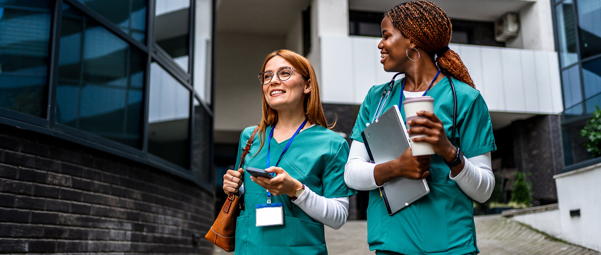 Two women dressed in medical scrubs, walking side by side outside a modern building. One woman is holding a smartphone and a leather bag, while the other is holding a cup of coffee and a laptop. They both appear to be smiling and engaged in conversation.