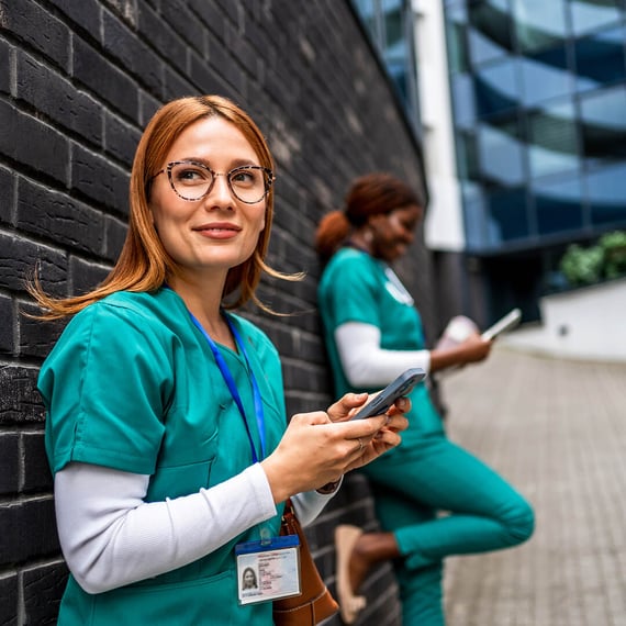 Zwei medizinische Fachkräfte, beide in grüner OP-Kleidung, stehen draußen vor einer schwarzen Backsteinmauer. Die Frau im Vordergrund hält ein Smartphone in den Händen, lächelt und blickt in die Kamera. Sie hat langes rotes Haar und trägt eine Brille; um den Hals trägt sie ein Namensschild. Im Hintergrund steht etwas weiter entfernt eine weitere Person, offenbar eine Kollegin, die ebenfalls ein Mobilgerät in der Hand hält. Die Kulisse wirkt modern und professionell; hinter den beiden ist ein großes Gebäude zu sehen.