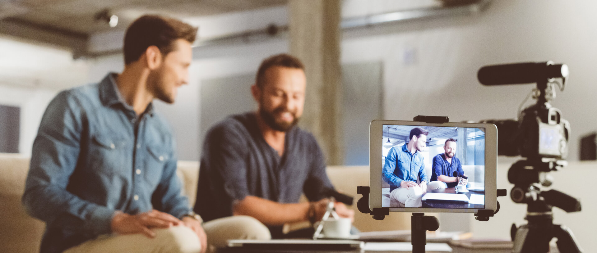 Zwei Männer sitzen in einer entspannten Umgebung auf einem Sofa und unterhalten sich oder führen ein Interview. Sie sind leger gekleidet; einer trägt ein Jeanshemd, der andere ein dunkelblaues T-Shirt. Vor ihnen steht eine Kameraausrüstung: Eine Kamera ist auf sie gerichtet, die andere – ein Tablet – zeigt die Live-Ansicht der beiden Männer aus der Perspektive der Kamera. Der Hintergrund ist ein moderner Raum im Industriestil mit freiliegenden Decken und sanfter Beleuchtung, der eine ungezwungene und dennoch professionelle Atmosphäre für die Aufzeichnung schafft.