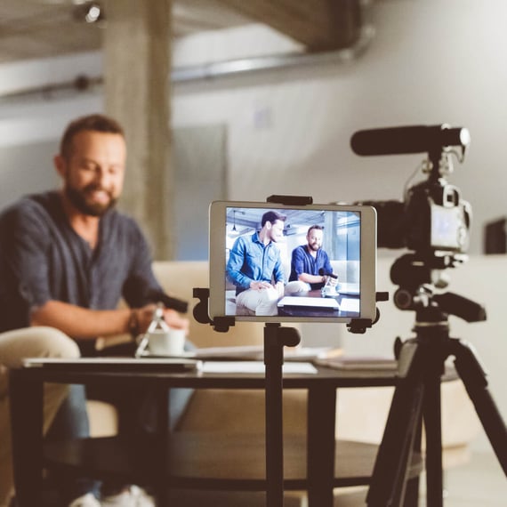 A close-up shot of a tablet displaying a live view of two men sitting across from each other at a table, having a conversation. The men are casually dressed, with one wearing a denim shirt and the other a dark blue T-shirt. The tablet is set up on a tripod, and a camera with a microphone is positioned nearby, capturing the scene. The background shows an industrial-style room with soft lighting, emphasizing the professional yet casual setting for the recording. The focus is on the live view of the two men on the tablet screen, with a blurred foreground of the camera setup.