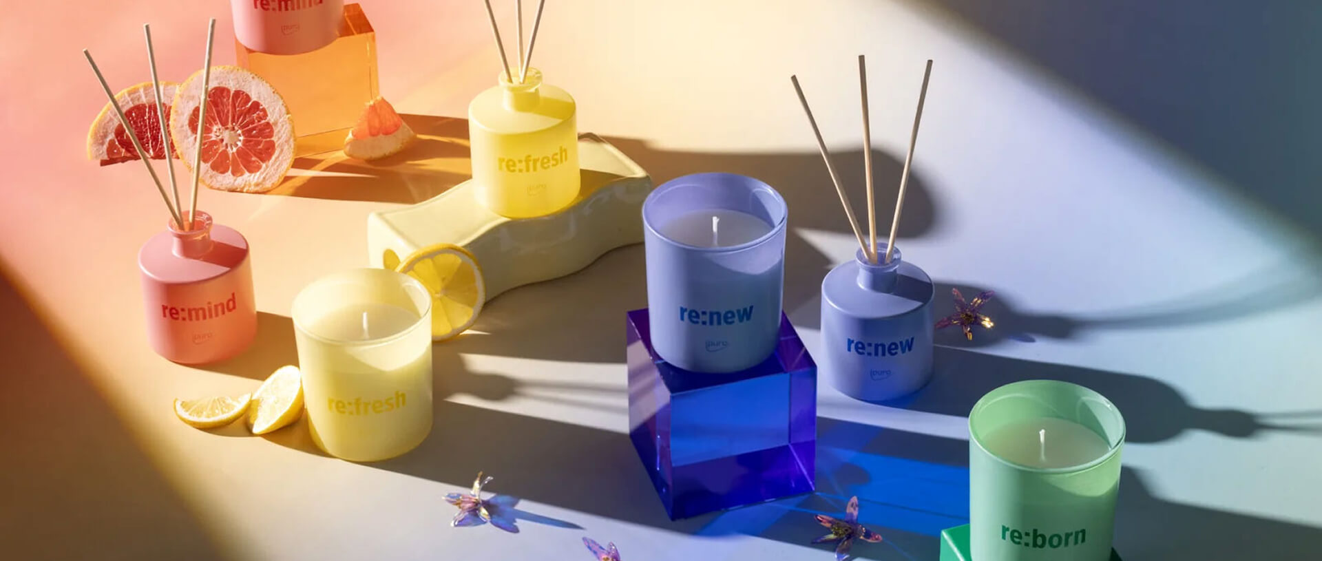 Photo showcasing a selection of home fragrances and candles arranged by colors: pink, yellow, purple and green. There are dried fruits and flowers scattered between the products.