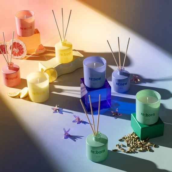 Foto, das eine Auswahl an Raumdüften und Kerzen zeigt, sortiert nach Farben: Rosa, Gelb, Violett und Grün. Zwischen den Produkten sind getrocknete Früchte und Blumen verstreut.