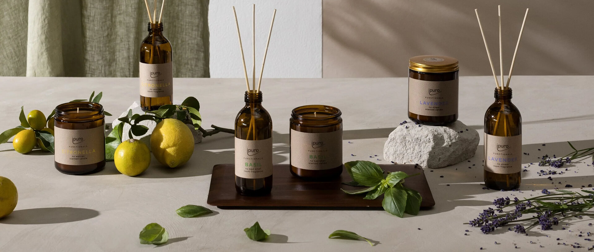 An elegant arrangement of amber glass containers with various scents, including citronella, basil, and lavender, from the brand iPuro. The scene features candles and reed diffusers surrounded by fresh ingredients like basil leaves, lavender sprigs, and lemons, all placed on a light stone surface. The neutral background and minimalistic design highlight the natural scents and soothing atmosphere.