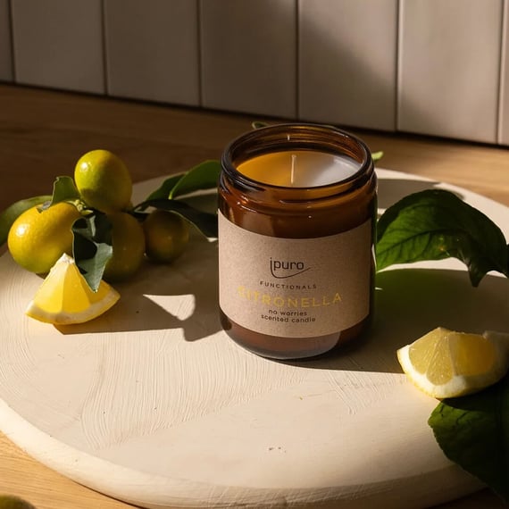 A close-up of a citronella-scented candle from iPuro placed on a round, textured wooden surface. The amber glass jar with a beige label is surrounded by fresh green leaves and yellow lemons, creating a natural and refreshing atmosphere. The warm sunlight highlights the scent of citronella, offering a calm and inviting ambiance.