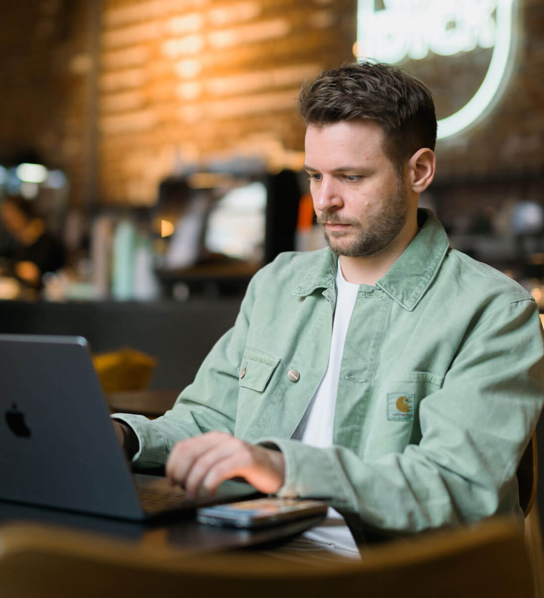 A man in a light green jacket works on a laptop at a wooden table in a warmly lit café. A smartphone lies beside the laptop, while the softly blurred background shows exposed brick walls, shelves and illuminated signage. | Business platform SUNZINET