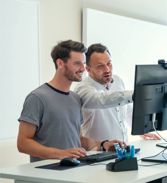 Zwei Männer stehen gemeinsam an einem Schreibtisch vor einem Computerbildschirm. Der Mann links trägt ein gestreiftes T-Shirt und bedient die Tastatur, während der Mann rechts in einem weißen Hemd etwas auf dem Monitor zeigt. Beide wirken konzentriert und engagiert, als ob sie gemeinsam an einem Projekt arbeiten oder etwas besprechen. 