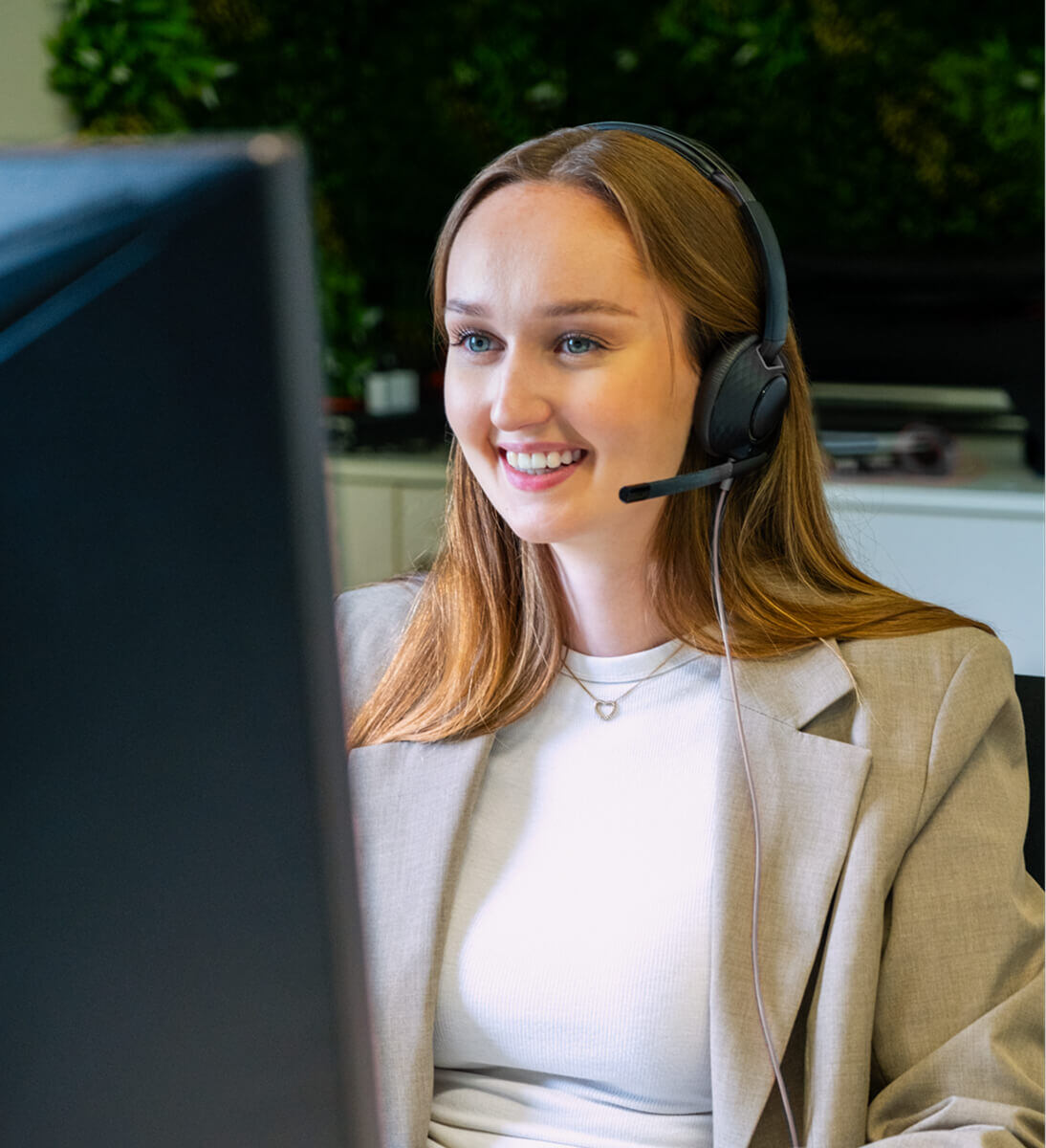 Eine junge Frau mit Headset sitzt an einem Schreibtisch vor einem Bildschirm und lächelt. Sie trägt ein helles Oberteil und ein beigefarbenes Sakko. Die Umgebung wirkt wie ein modernes Büro mit grüner Pflanzendekoration im Hintergrund. Die Szene vermittelt den Eindruck eines freundlichen Kundengesprächs oder virtuellen Meetings.