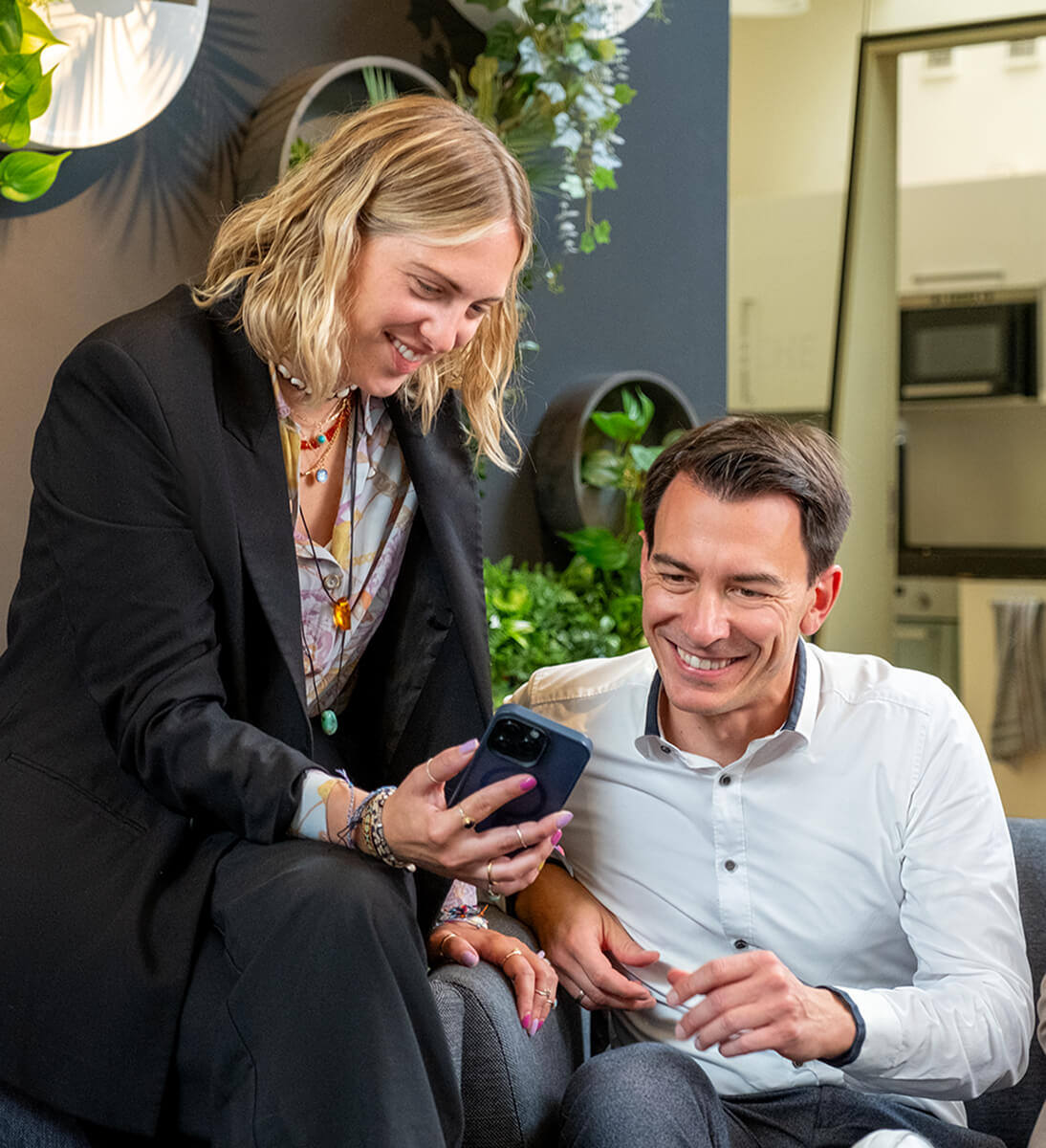 Eine Frau zeigt einem Mann, der neben ihr auf dem Sofa sitzt, etwas auf ihrem Mobiltelefon. Alle lächeln und sind in ein freundliches Gespräch vertieft. Die Umgebung ist modern mit viel Grün im Hintergrund | Interne Kommunikation SUNZINET