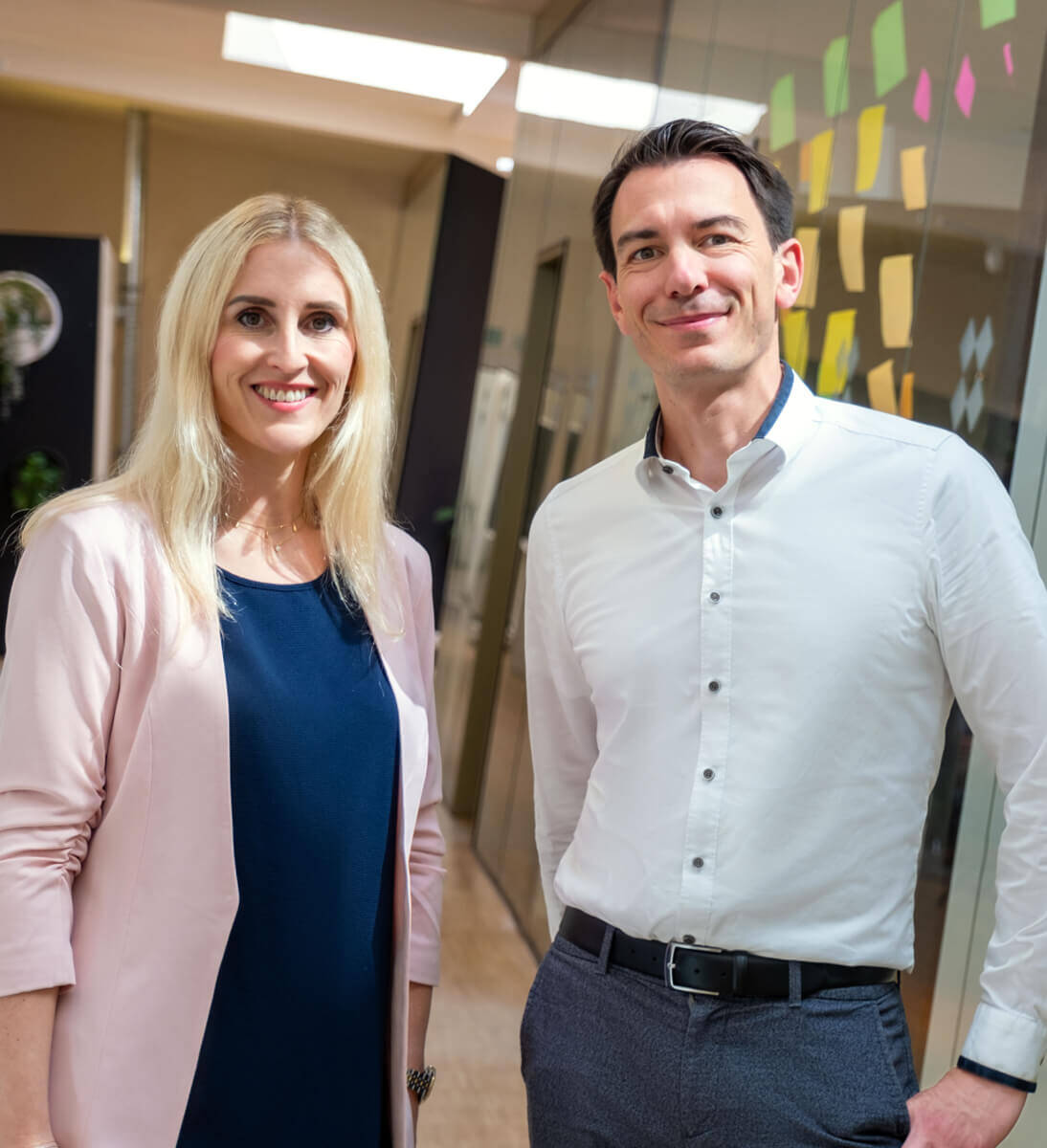 Two professionals standing together in a modern office environment, smiling confidently. The woman, with long blonde hair, is wearing a light pink jacket over a blue dress. The man, with short dark hair, is dressed in a white shirt and gray pants. The background shows a wall decorated with colorful sticky notes, indicating a collaborative work environment. | Market entry framework SUNZINET