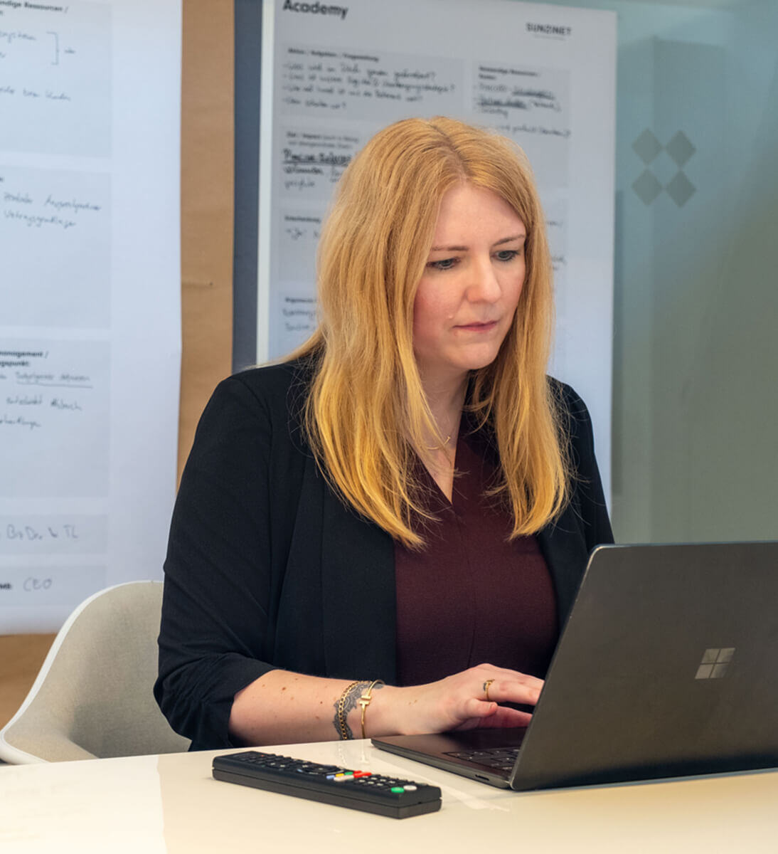 Eine Frau arbeitet an einem Laptop an einem Tisch, mit einem Whiteboard und Notizen im Hintergrund. Sie ist auf ihre Arbeit konzentriert, trägt ein kastanienbraunes Hemd und hat lange blonde Haare | Interne Kommunikation SUNZINET