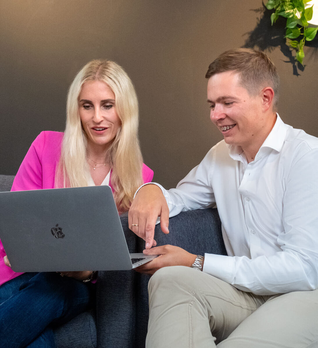 Eine Frau und ein Mann sitzen zusammen auf einem Sofa und schauen auf einen Laptop-Bildschirm. Die Frau trägt einen rosa Blazer, der Mann ein weißes Hemd. Beide lächeln und sind in ein Gespräch vertieft. SUNZINETA Mann und Frau arbeiten gemeinsam an einem Computerbildschirm, lächeln und sind mit der Aufgabe beschäftigt. Der Mann trägt ein rot-weiß gestreiftes Hemd, die Frau hat kurze blonde Haare und trägt ein weißes Hemd mit einer Halskette | Interne Kommunikation SUNZINET
