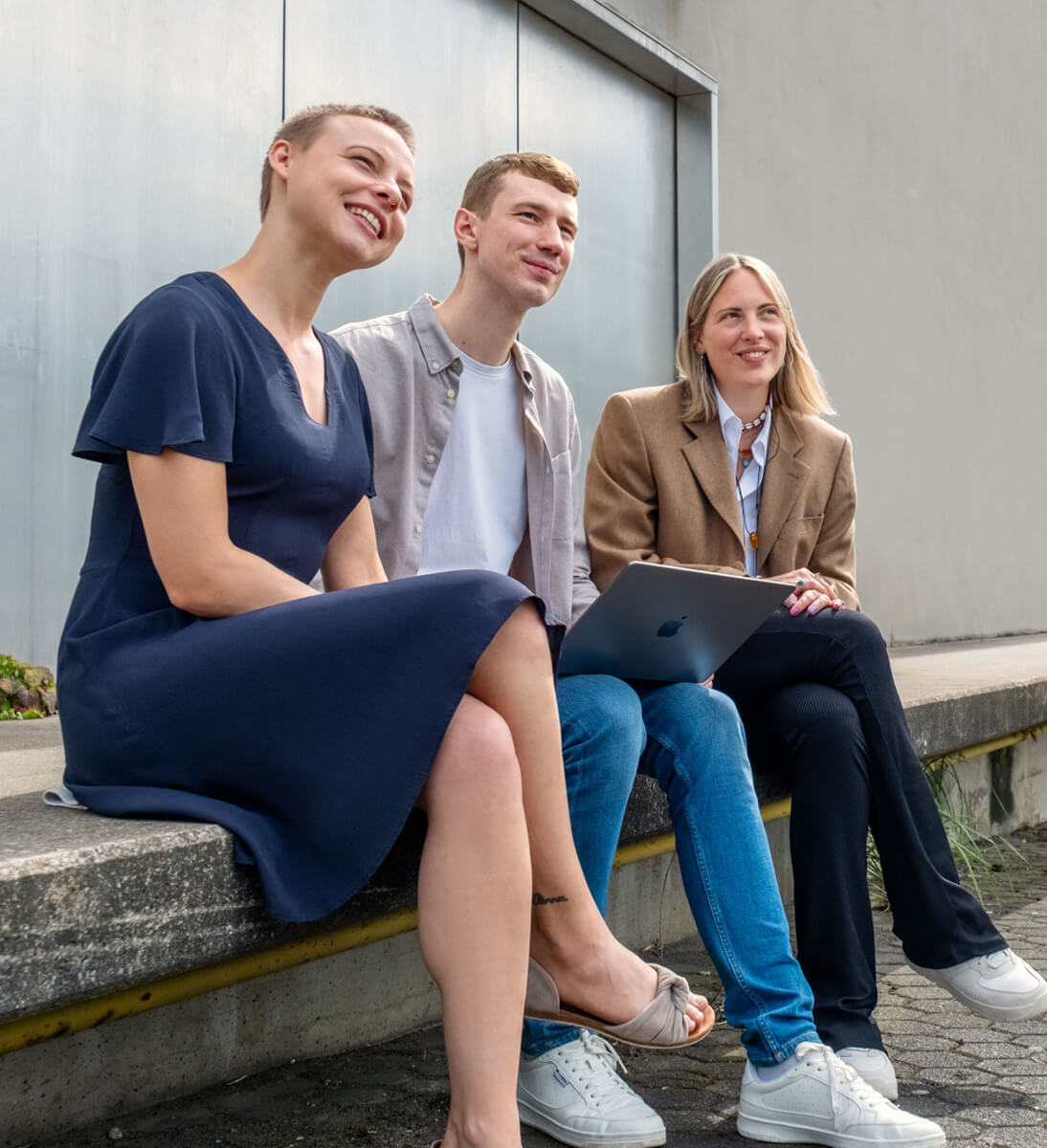 Drei junge Menschen sitzen draußen auf einer Treppe, lächeln und schauen in dieselbe Richtung, einer hält ein Laptop. | KI-Hub von SUNZINET