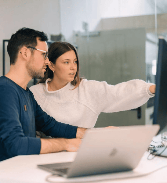 Eine Frau und ein Mann sitzen gemeinsam an einem Schreibtisch vor einem Computerbildschirm. Die Frau zeigt mit ausgestrecktem Arm auf den Monitor und erklärt etwas, während der Mann aufmerksam zuhört. Auf dem Tisch steht ein aufgeklappter Laptop. Die Szene wirkt wie eine konzentrierte Zusammenarbeit in einem modernen Büro.