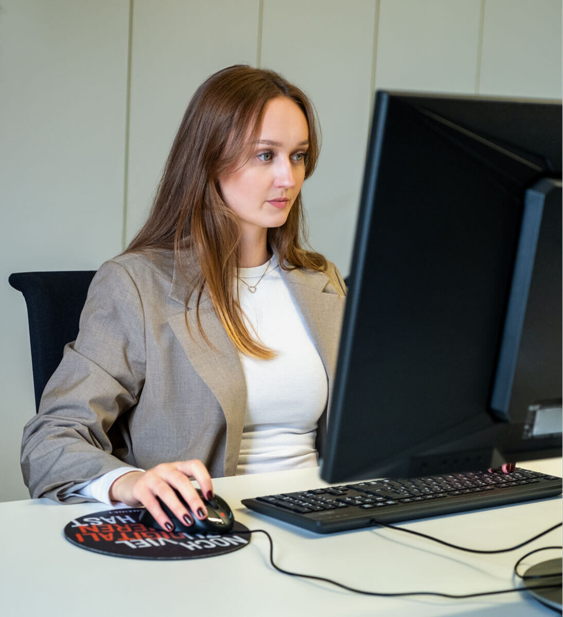 Eine junge Frau mit langen braunen Haaren sitzt konzentriert an einem Schreibtisch und arbeitet an einem großen Monitor. Sie trägt einen beigen Blazer über einem weißen Oberteil und steuert mit der Maus die Benutzeroberfläche auf dem Bildschirm. Die Umgebung wirkt professionell und modern, passend zum Thema 