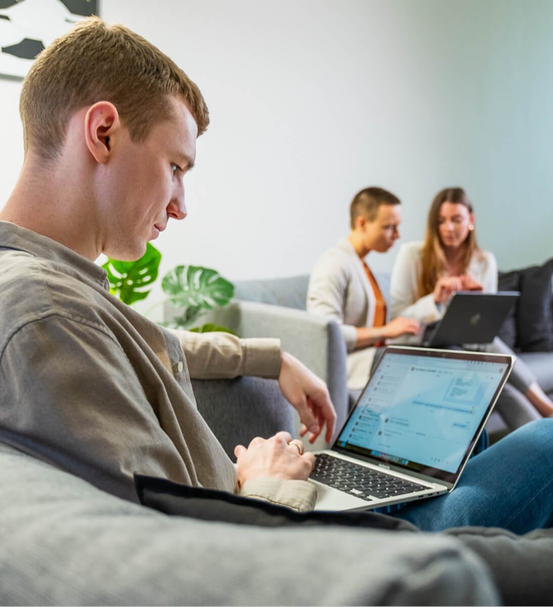 Ein junger Mann sitzt entspannt auf einem grauen Sofa und arbeitet konzentriert an einem Laptop. Im Hintergrund befinden sich zwei weitere Personen auf einem anderen Sofa, die ebenfalls mit Laptops arbeiten und sich austauschen. Die Szene zeigt eine moderne, kollaborative Arbeitsumgebung mit einer gemütlichen Atmosphäre, unterstrichen durch grüne Pflanzen im Hintergrund. Das Bild symbolisiert das Thema 
