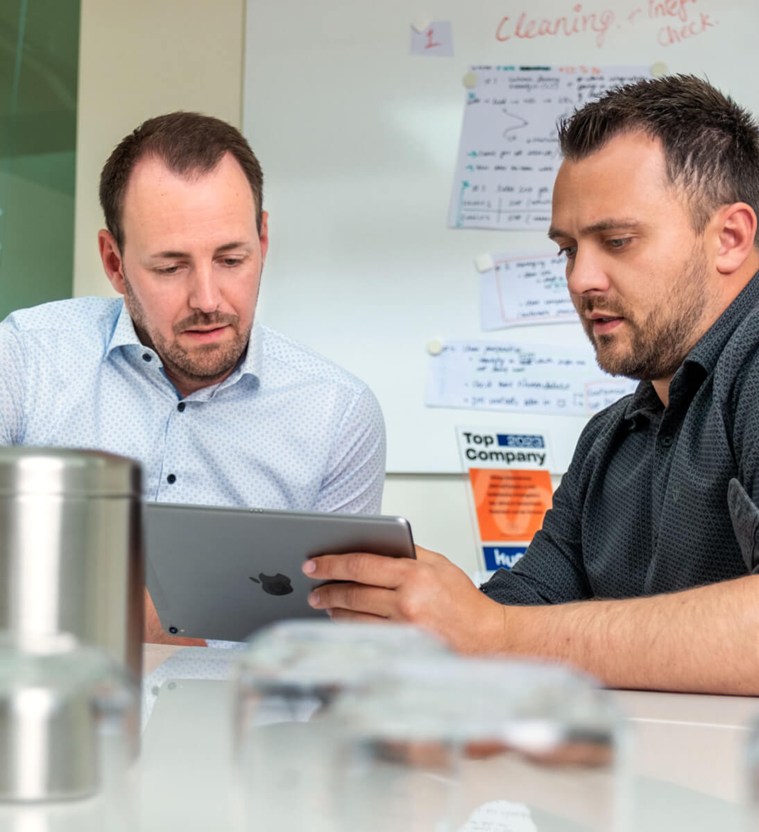 Zwei Männer sitzen an einem Tisch in einem modernen Büro und betrachten gemeinsam ein Tablet. Einer trägt ein helles Hemd, der andere ein dunkles Hemd mit hochgekrempelten Ärmeln. Im Hintergrund ist ein Whiteboard mit handschriftlichen Notizen und Plänen zu sehen. Auf dem Tisch stehen mehrere Trinkgefäße. Die Szene vermittelt eine konzentrierte Arbeitsatmosphäre und symbolisiert die Analyse der Integrationsmöglichkeiten für eine KI-Suchlösung.