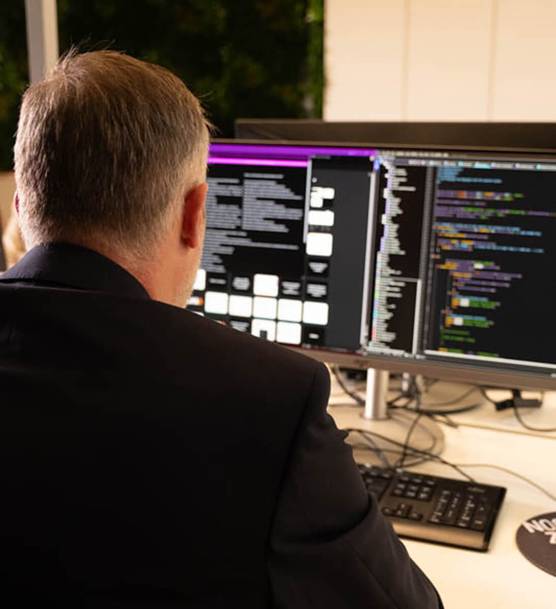 Ein Mann in einem dunklen Anzug sitzt an einem Schreibtisch und arbeitet an zwei großen Monitoren. Auf den Bildschirmen sind Codezeilen und eine Benutzeroberfläche mit strukturierten Texten zu sehen. Die Umgebung wirkt professionell und technisch, mit einem Fokus auf die Optimierung von Sprachmodellen und Embeddings für eine KI-Suchlösung. Im Hintergrund ist eine moderne Büroatmosphäre mit grünen Pflanzen sichtbar.