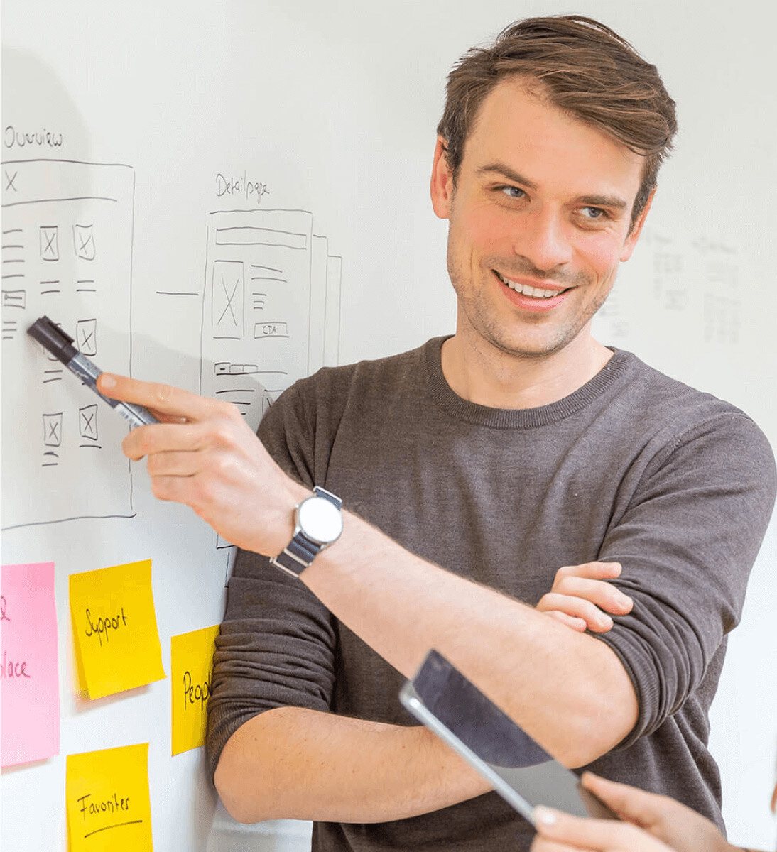 Ein lächelnder Mann mit braunen Haaren und einer dunklen Armbanduhr steht vor einem Whiteboard und zeigt mit einem schwarzen Marker auf eine Skizze von Wireframes und Prozessschritten. Um ihn herum befinden sich mehrere bunte Haftnotizen mit Beschriftungen. Die Szene wirkt wie ein Workshop oder eine Planungssitzung zur Identifikation relevanter Daten für die Implementierung einer KI-Suche.
