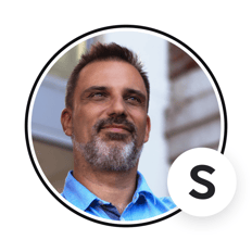 Portrait of a person with short dark hair and a salt-and-pepper beard, wearing a light blue shirt, displayed in a circular frame with an “S” badge overlay.